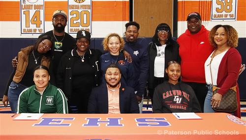 Group of people standing around three people that are seated at a long table. "EHS" is printed on the orange table cloth.